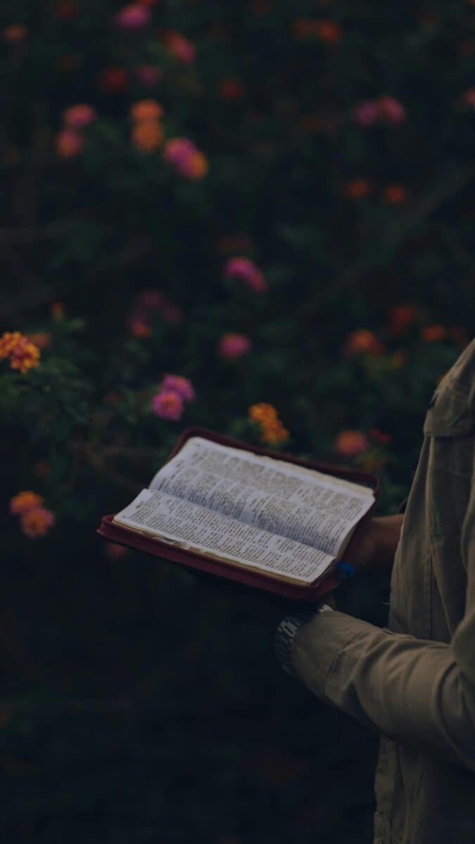 A close-up of a person holding an open book amidst vibrant flowers outdoors.